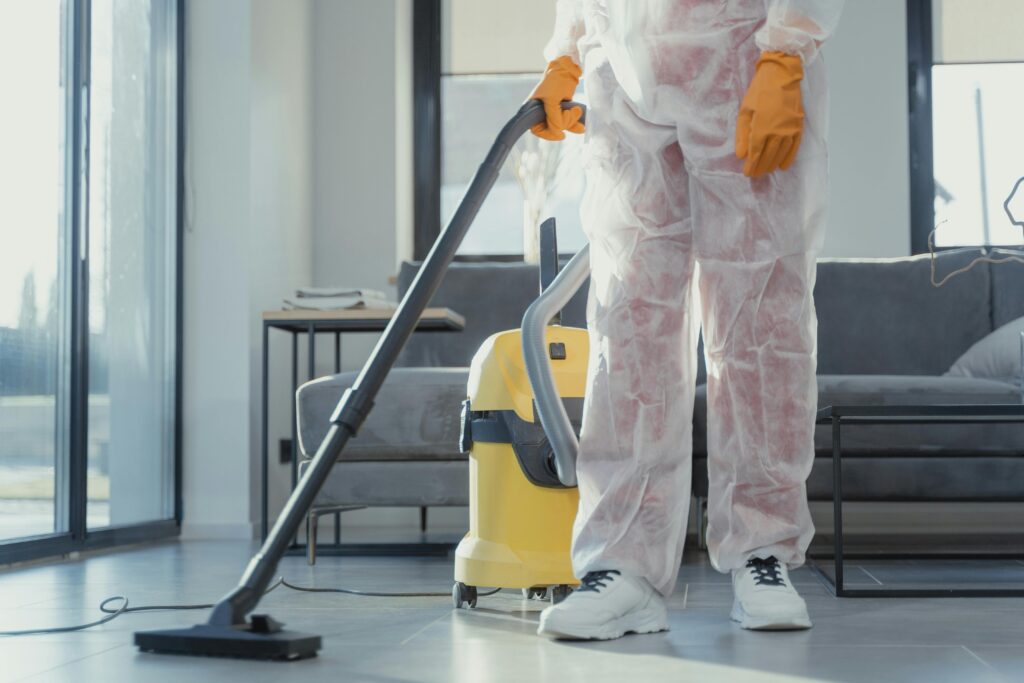 A cleaner in protective coveralls and gloves vacuuming a modern living room.